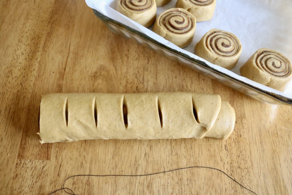 log of cinnamon roll dough being sliced with string and sliced cinnamon rolls on a baking sheet