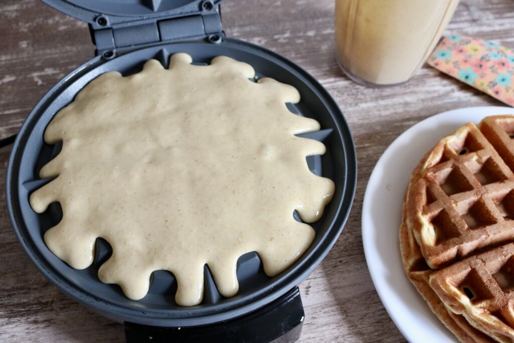 waffle batter on a waffle iron with a cooked waffle, waffle batter, and a spatula off to the right side