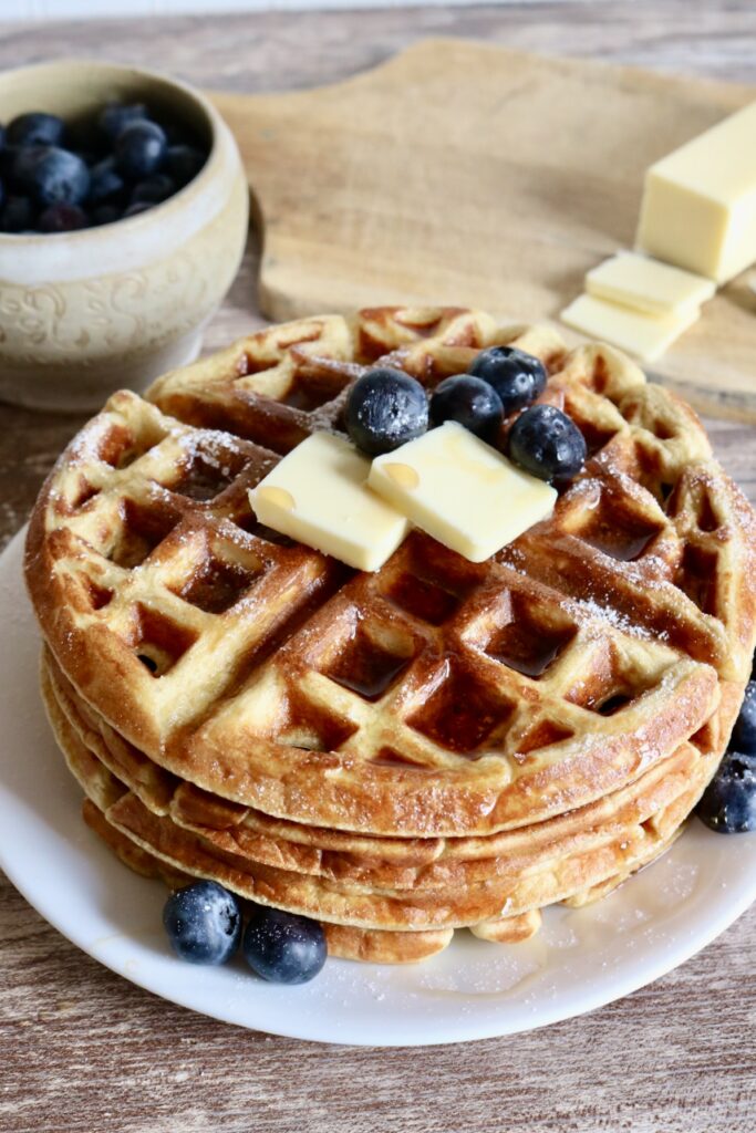 stack of four waffles on a plate with butter, blueberries, and syrup on top. a cup of blueberries and a stick of butter are behind it