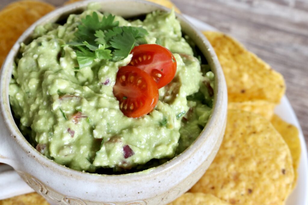 protein guacamole dip in a serving dish with fresh cilantro and a cherry tomato cut in half as a garnish. tortilla chips are on the side