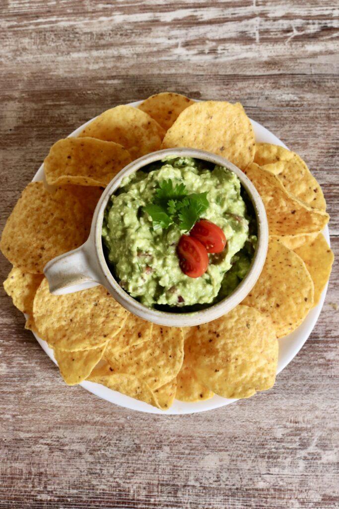 top view of guacamole in a serving dish on a white plate with tortilla chips around it