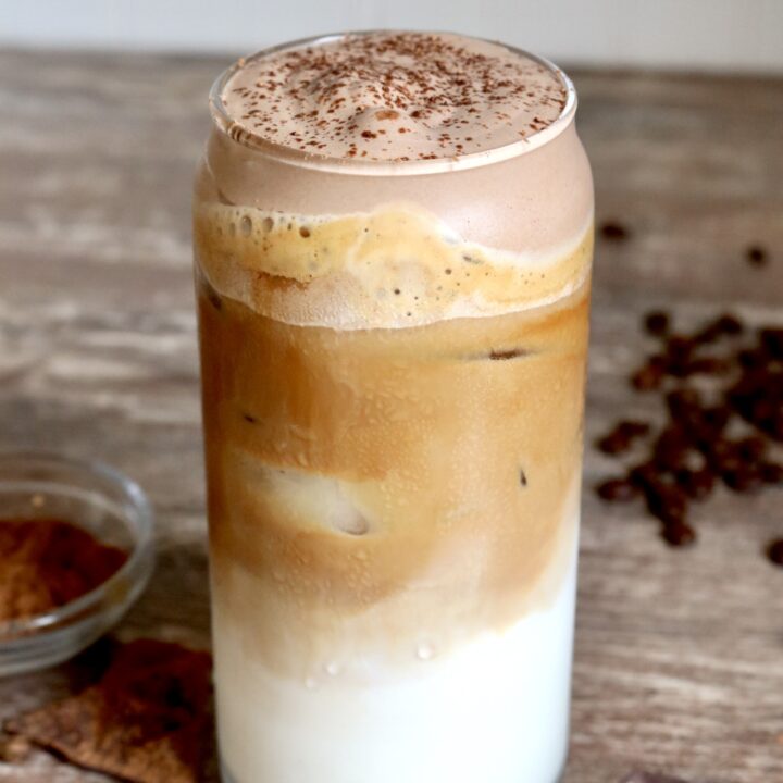 iced latte with chocolate cold foam coffee beans, chocolate bars, and cocoa powder scattered around