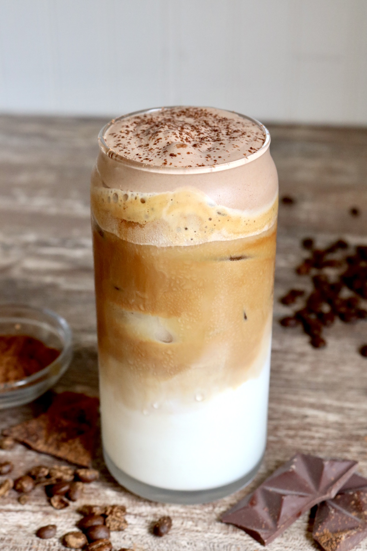 iced latte with chocolate cold foam coffee beans, chocolate bars, and cocoa powder scattered around