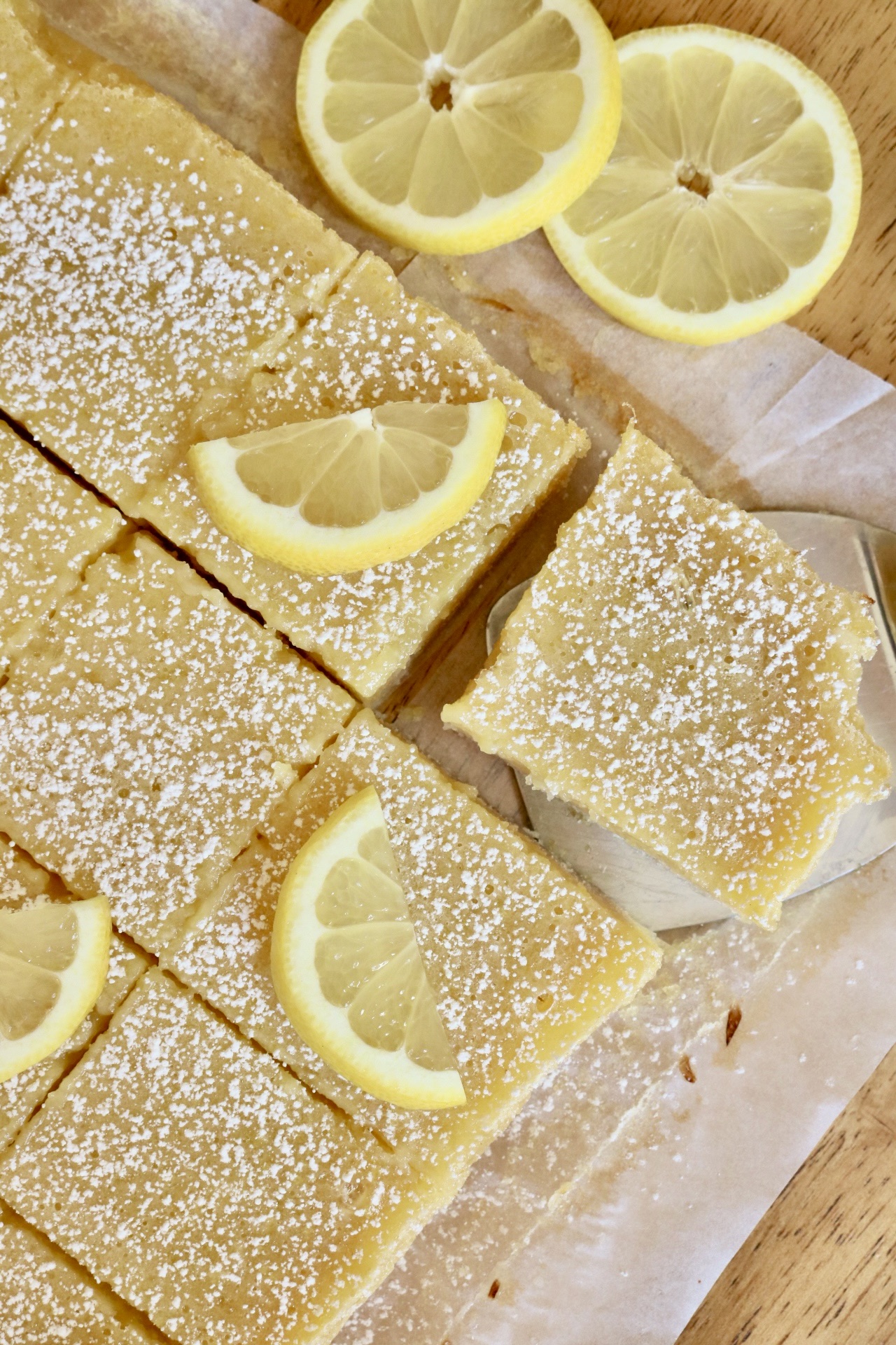 lemon bar squares with lemon round next to it and a small spatula under the corner bar