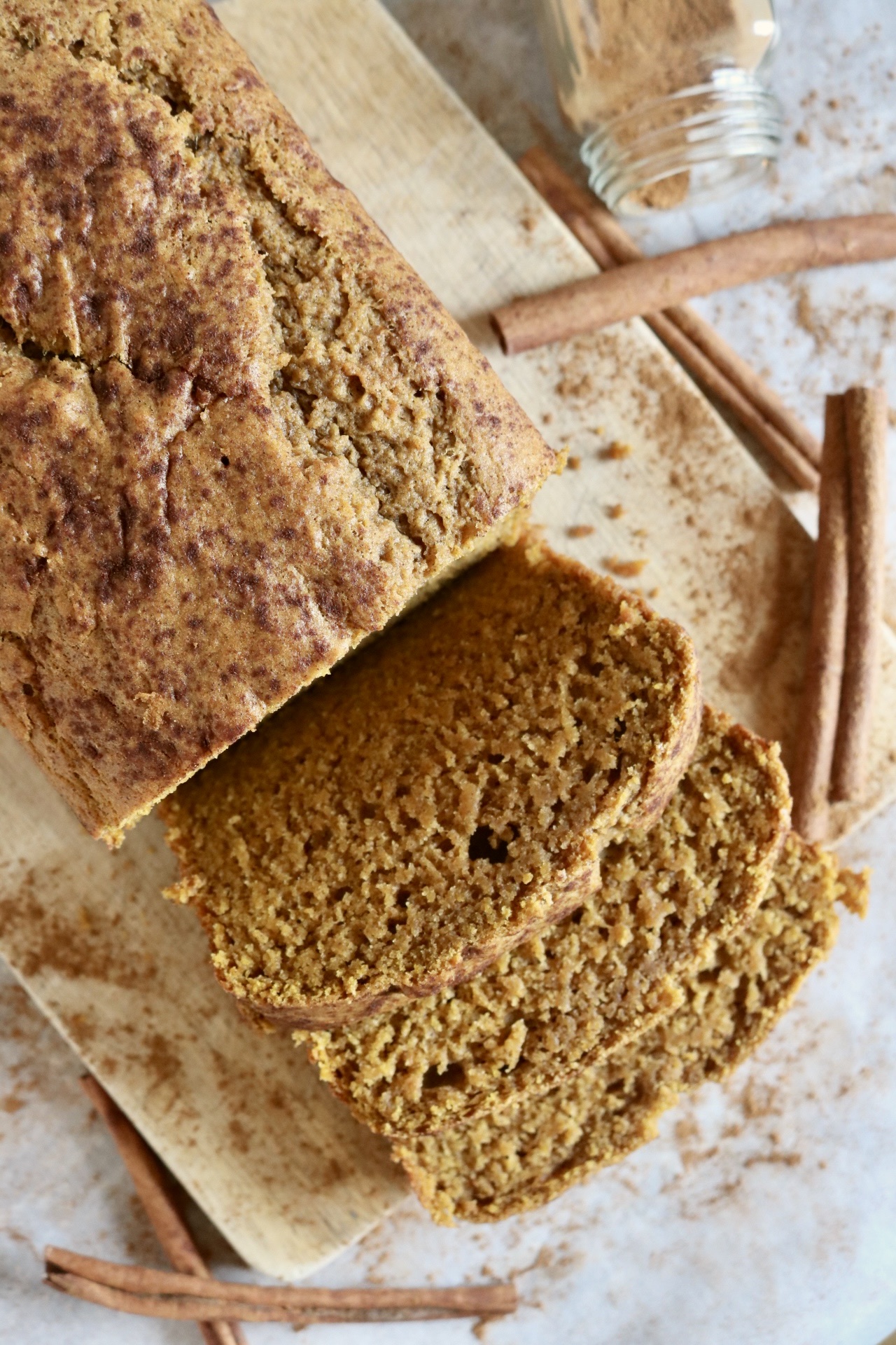 sliced loaf of pumpkin bread on a wooden cutting board with cinnamon sticks and a glass jar of ground cinnamon on its side beside the loaf