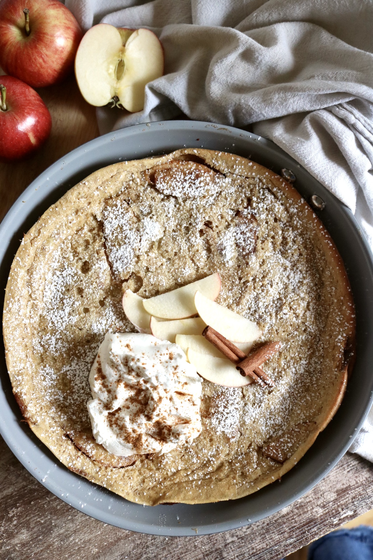 apple dutch baby pancake in a skillet with whipped cream cinnamon and sliced apples and cinnamon sticks on top