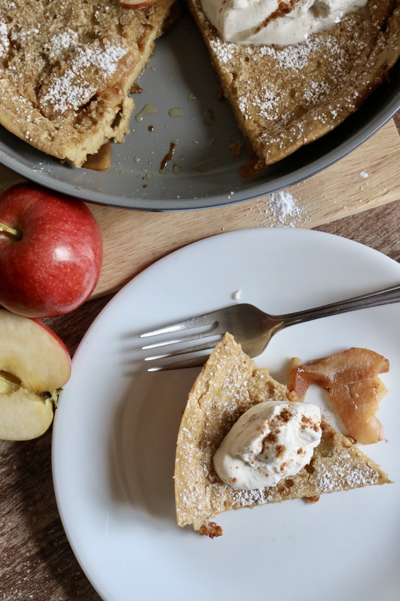 piece of apple dutch baby on a white plate with a fork and whipped cream and cinnamon sprinkled on top with the pan of dutch baby pancakes on the side and fresh apples beside the plate