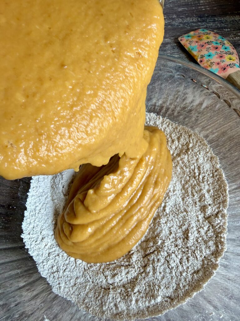 pouring wet pumpkin bread ingredients into a flour mixture a pink spatula is off to the side